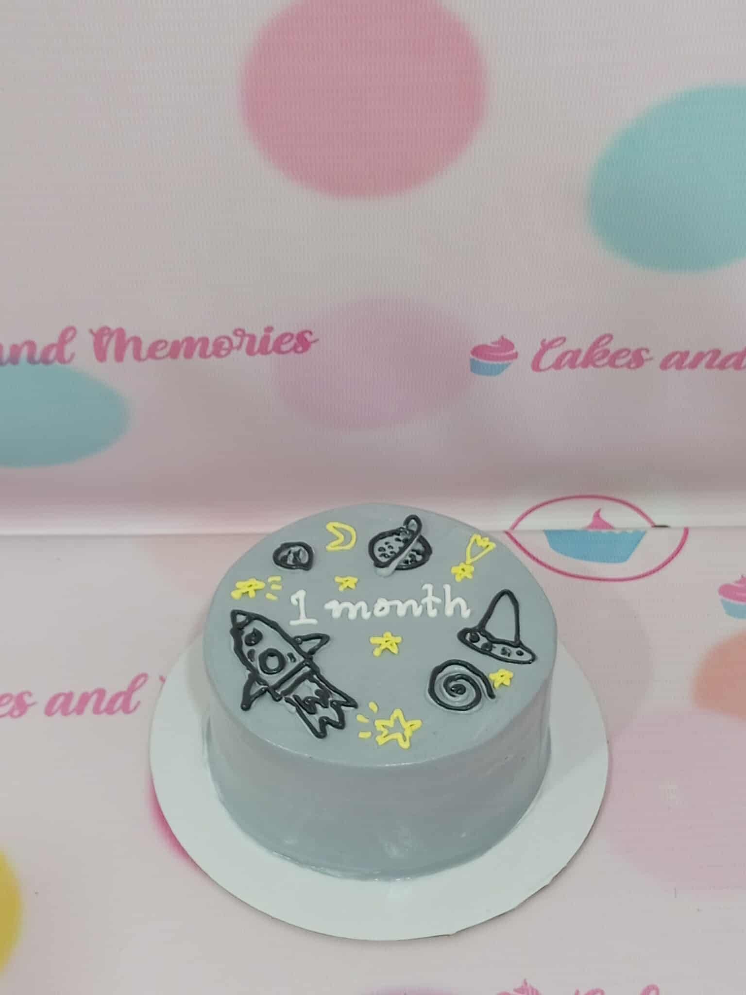 One-tier grey space cake with hand-piped rocket, UFO, planets, yellow stars, and '1 month' text in buttercream.