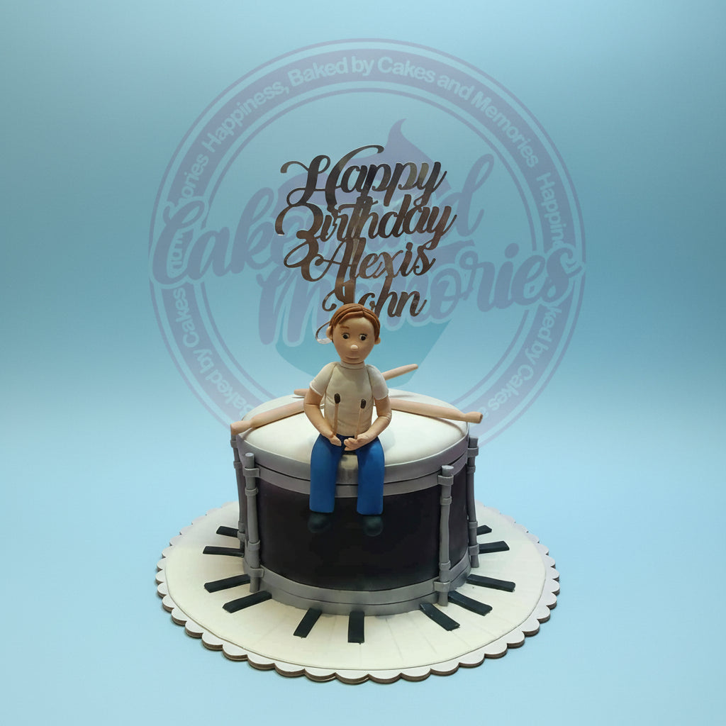 One-tier custom fondant birthday cake with a drum design, a boy drummer figure, piano key board, and 'Happy Birthday Alexis John' topper. Music cake.