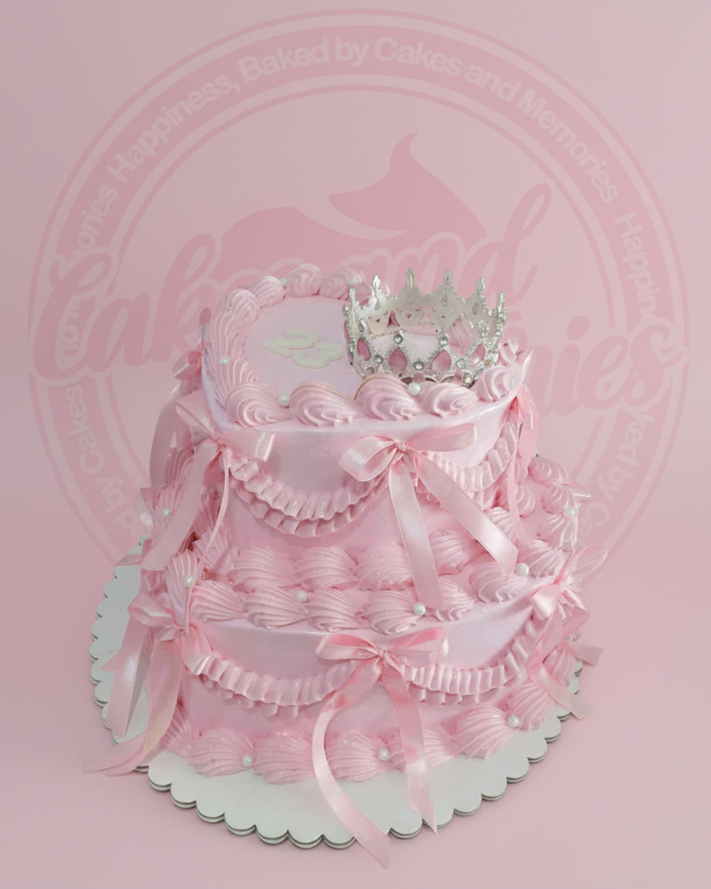 Two-tier pink princess crown cake with ruffled buttercream, satin ribbons, edible pearls, and a silver crown topper for a birthday.