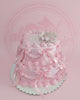 Two-tier pink princess crown cake with ruffled buttercream, satin ribbons, edible pearls, and a silver crown topper for a birthday.