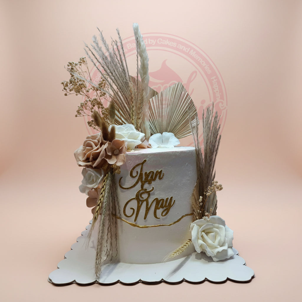Custom wedding cake with boho dried florals, sugar roses, and "Juan & May" topper in white, gold, and earthy tones.