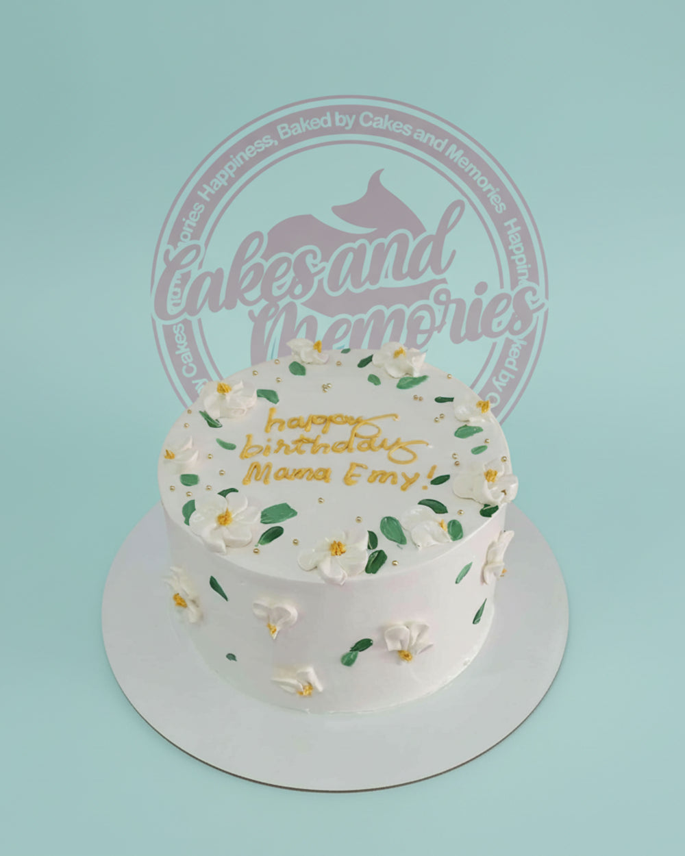 One-tier birthday cake with white buttercream, delicate white flowers, green leaves, gold pearls, and 'happy birthday Mama Emy!'