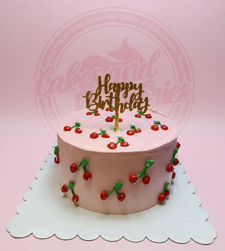 One-tier pink buttercream birthday cake with hand-piped red cherries and a gold 'Happy Birthday' topper on a scalloped board.