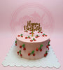 One-tier pink buttercream birthday cake with hand-piped red cherries and a gold 'Happy Birthday' topper on a scalloped board.