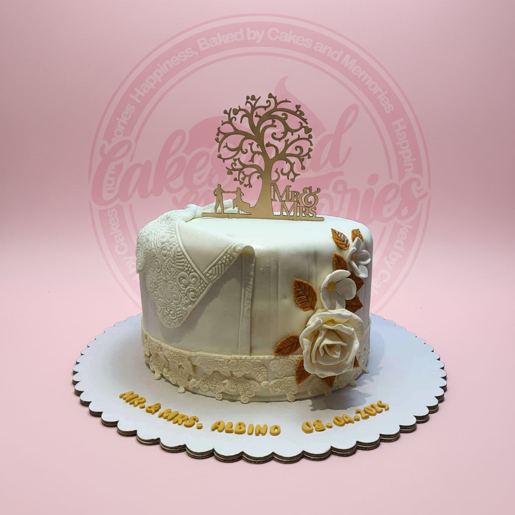 White fondant wedding cake with elegant lace details, cream roses, and a golden 'Mr & Mrs' tree topper. Personalized with names and date.