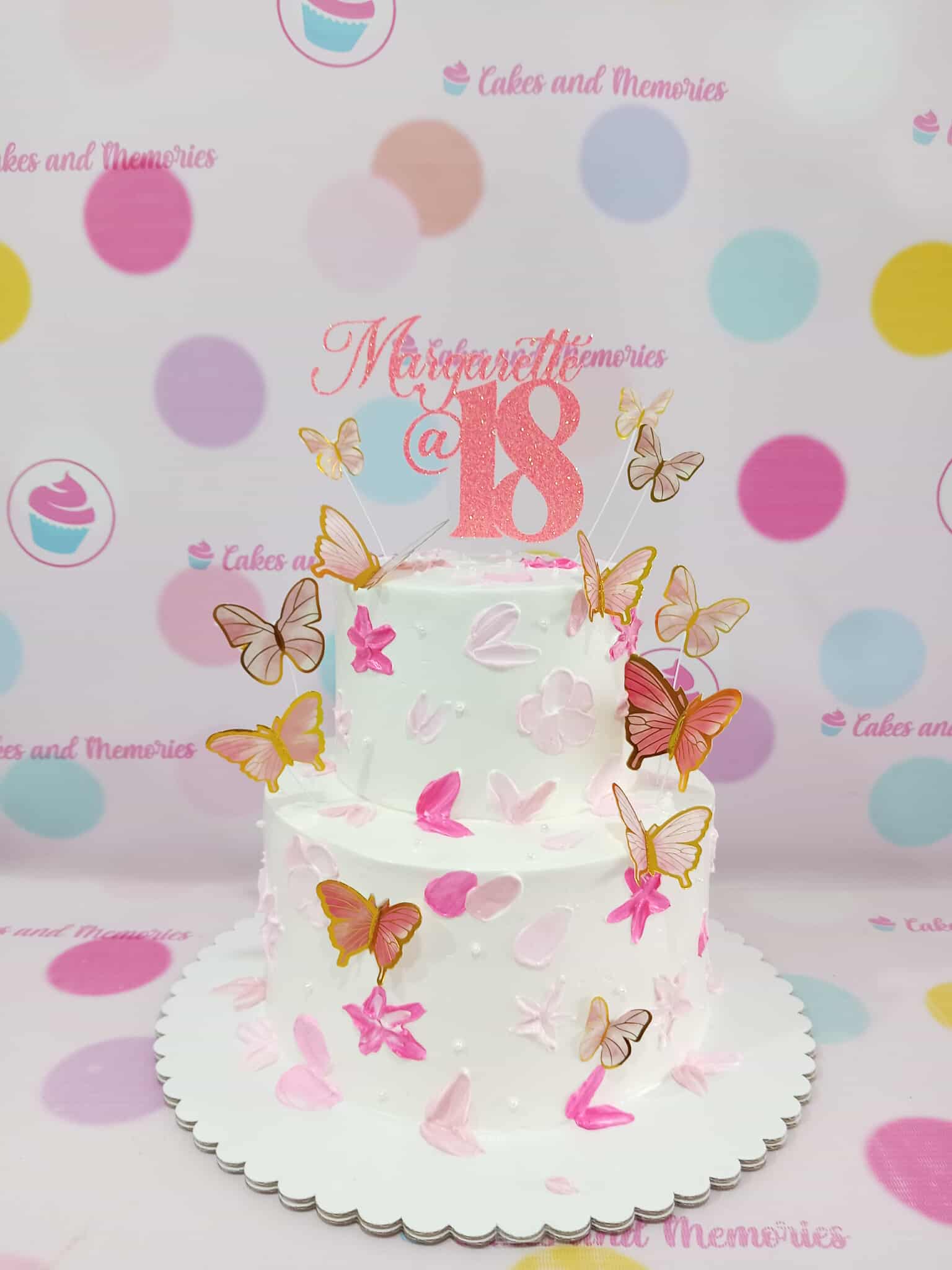 Two-tier custom birthday cake with white buttercream, pink strokes, glitter 'Margarett@ 18' topper, and fluttering pink & gold butterflies.