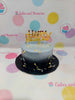 One-tier round birthday cake with ombre grey to black buttercream, topped with a gold 'Happy Birthday' topper, silver pearls, and metallic foil flakes.