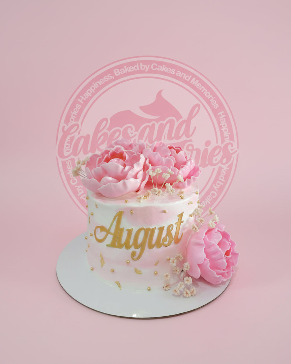 One-tier round floral cake with pink and white watercolor icing, gold 'August' text, and pink peonies.