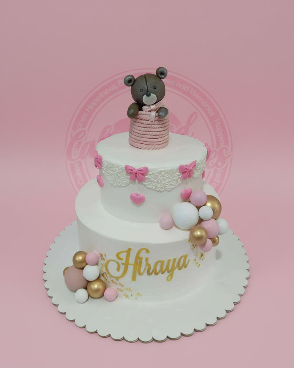 Two-tier teddy bear fondant cake with pink bows, hearts, gold "Hiraya" name, and metallic gold, pink, white spheres.