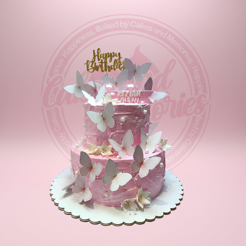 Two-tier pink butterfly birthday cake with white pearls, edible flowers, and a gold 'Happy Birthday' topper for Keziah Aishi.