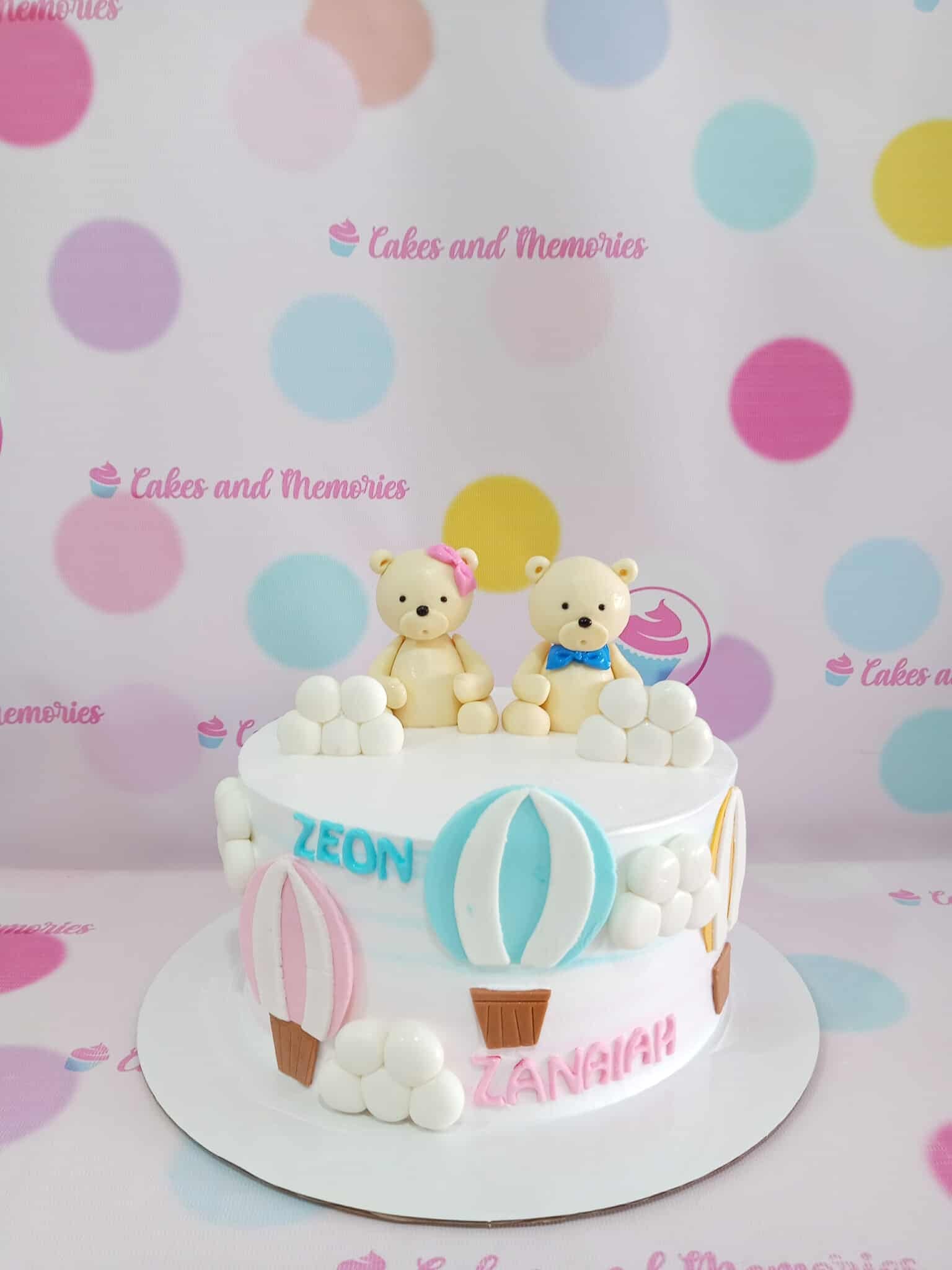 One-tier hot air balloon & teddy bear custom cake with fondant details, names Zeon & Zanaian, in white, pink, blue, yellow.