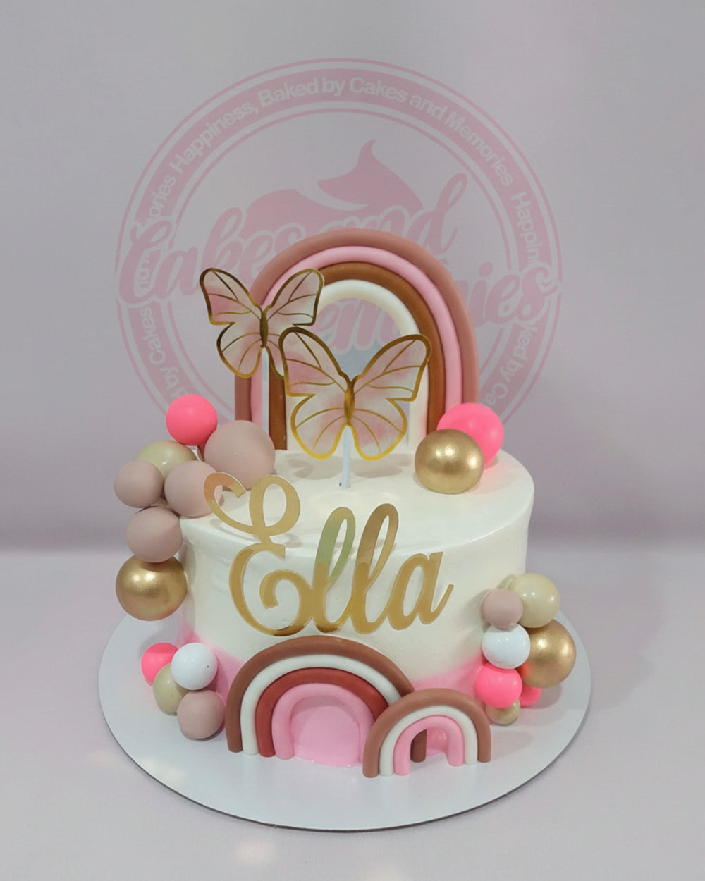 One-tier white buttercream boho rainbow birthday cake with gold 'Ella' name, pink, gold, and cream spheres, and delicate butterfly toppers.