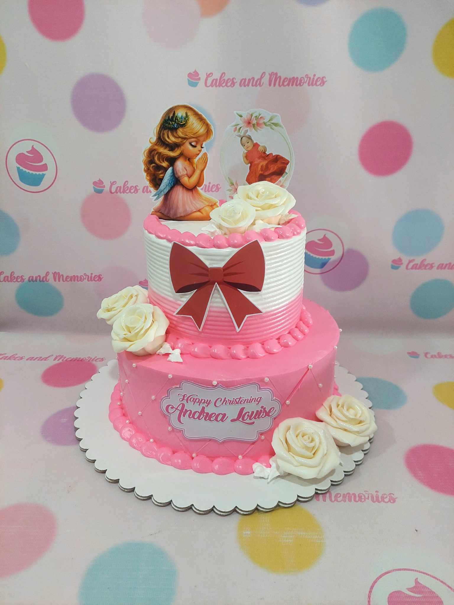Two-tier christening cake with praying angel and baby toppers, personalized plaque, and white roses in pink and white.