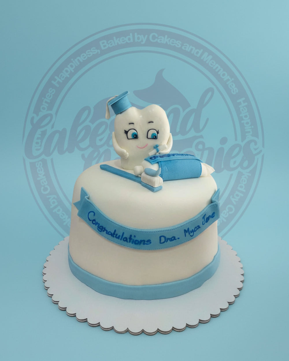 One-tier fondant dental graduation cake: cute tooth in cap, toothbrush, toothpaste, and "Congratulations Dra. Myca Jene" banner, white and blue.