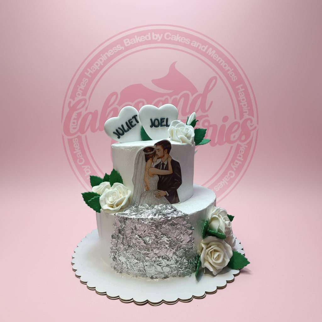 Two-tier wedding cake with kissing bride and groom edible print, 'JULIET' & 'JOEL' heart toppers, white roses, and silver foil.