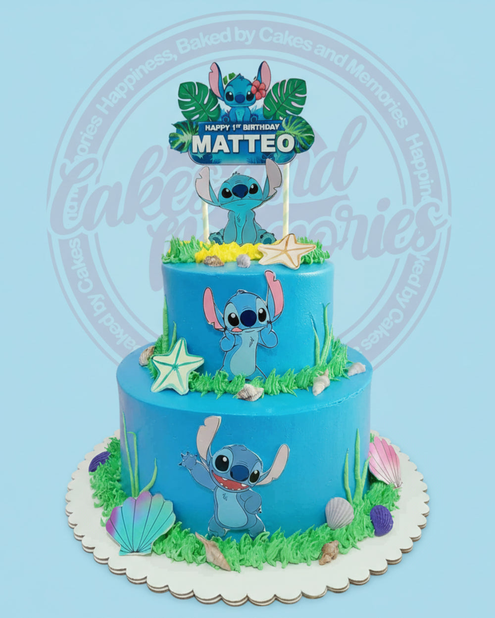 Two-tier Lilo & Stitch cake with blue icing, Stitch cutouts, seashells, starfish, and green grass, featuring 'HAPPY 1ST BIRTHDAY MATTEO'.
