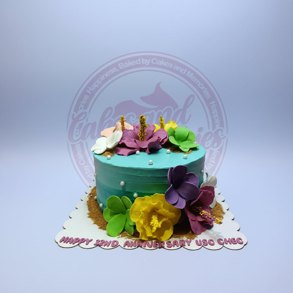 One-tier tropical anniversary cake with ombre blue-green buttercream, vibrant sugar paste hibiscus flowers, sand decor, and 'Happy 32nd Anniversary USC CHEC' text.