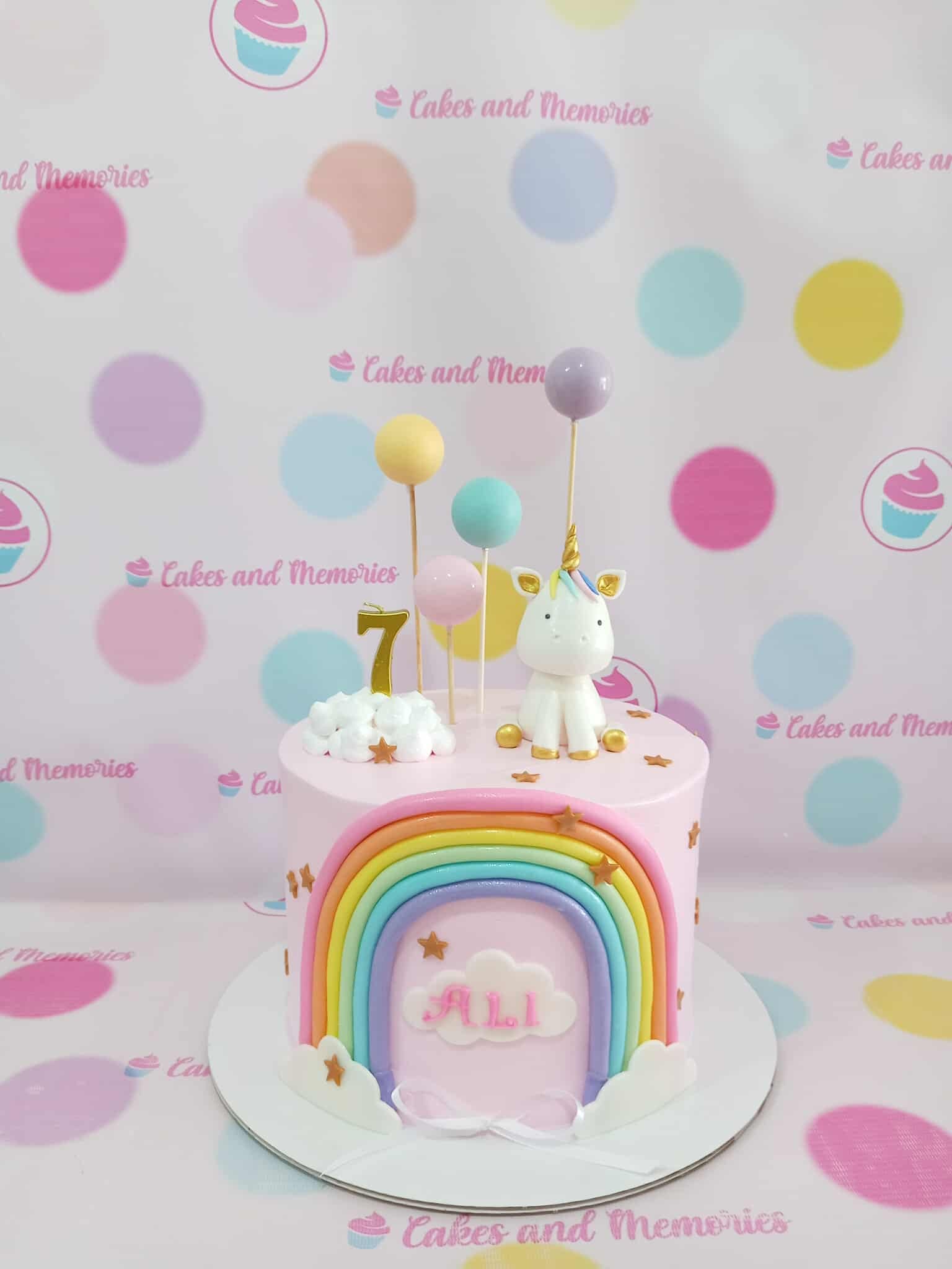 One-tier Unicorn Rainbow Birthday Cake with pastel rainbow, white unicorn, clouds, gold stars. Personalized with 'ALI' & '7'. Pink icing.