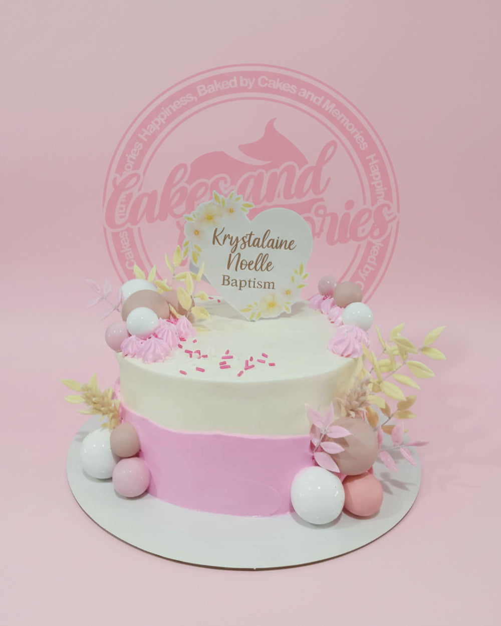 One-tier baptism cake with white and pink buttercream, adorned with edible spheres, meringues, foliage, and a 'Krystalaine Noelle Baptism' topper.
