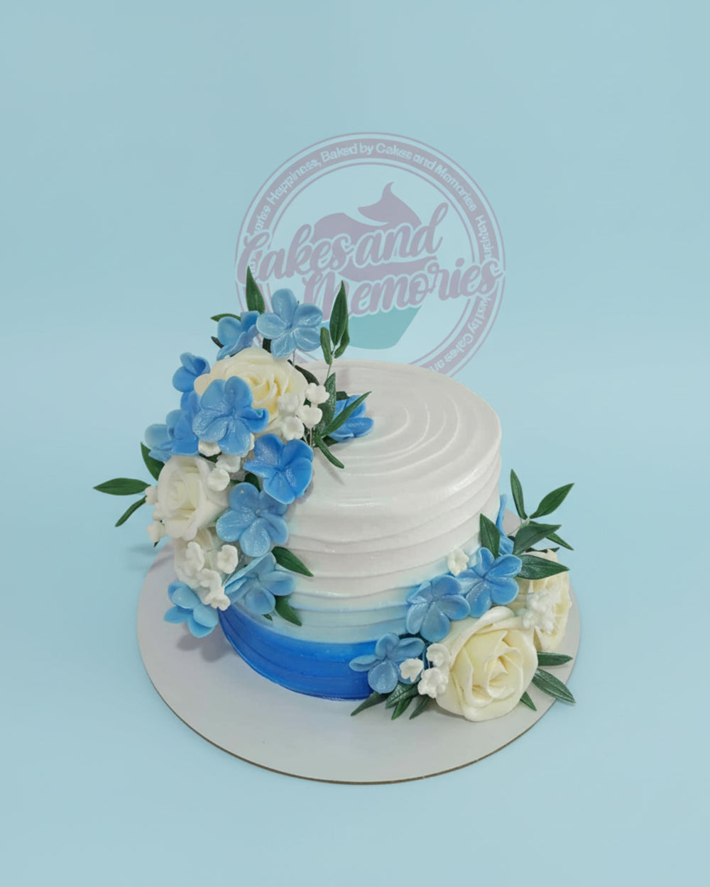 Custom buttercream cake with white to blue ombre icing, adorned with white roses and blue sugar flowers and green leaves.
