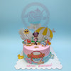 One-tier Minnie Mouse birthday cake with beach theme toppers, 'Amaia' plaque, pink & blue icing, and 'Happy Birthday!' text.