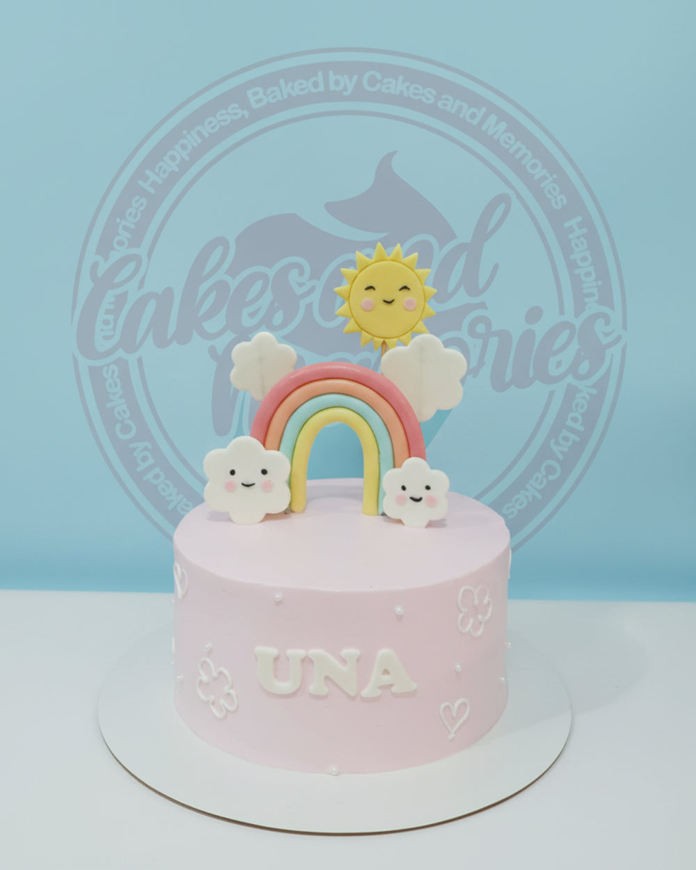 One-tier pink buttercream birthday cake with smiling rainbow, sun, and clouds, personalized with "UNA".