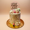 Elegant 1-tier buttercream birthday cake with gold leaf, piped details, pastel flowers, macarons, and a 'Happy 75th Birthday' topper.