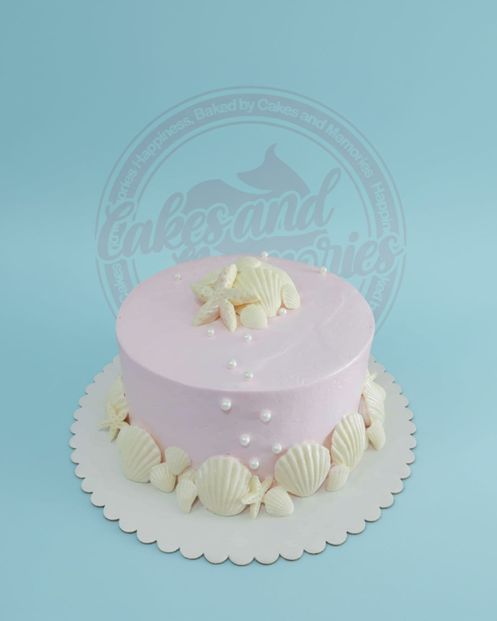 One-tier pink buttercream cake with white edible seashells, starfish, and pearls on a scalloped cake board, perfect for an under the sea celebration.