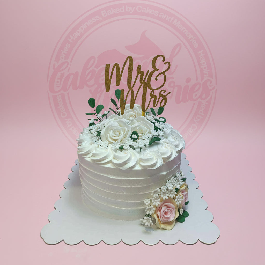 Elegant white floral wedding cake with a gold 'Mr & Mrs' topper, delicate white, blush, and cream sugar flowers, and green leaves.
