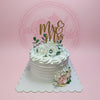 Elegant white floral wedding cake with a gold 'Mr & Mrs' topper, delicate white, blush, and cream sugar flowers, and green leaves.
