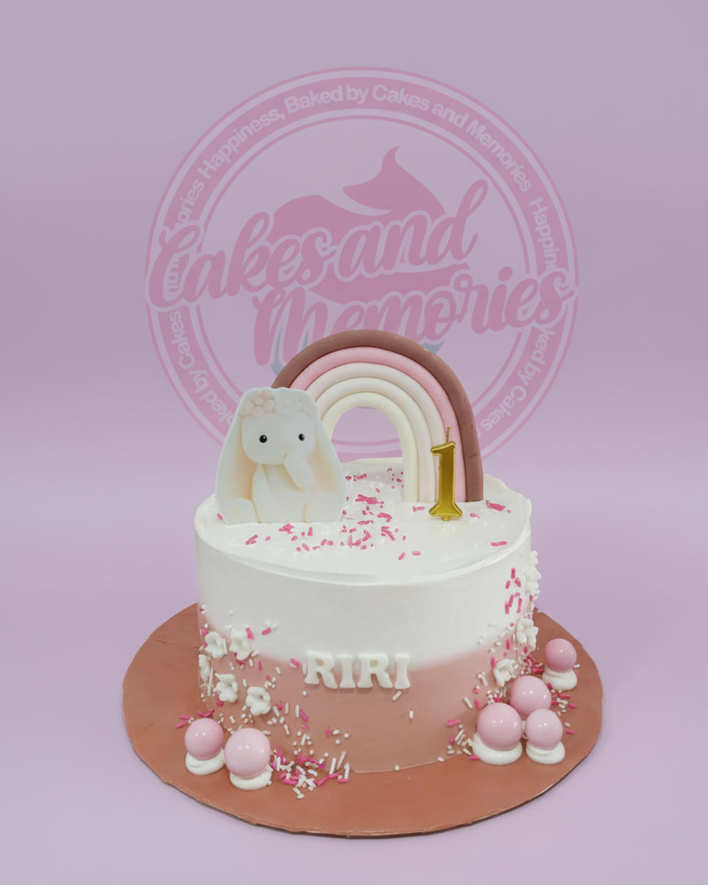 One-tier bunny and rainbow first birthday cake with ombre pink buttercream, 'RIRI' name, and '1' candle on a rose gold board.
