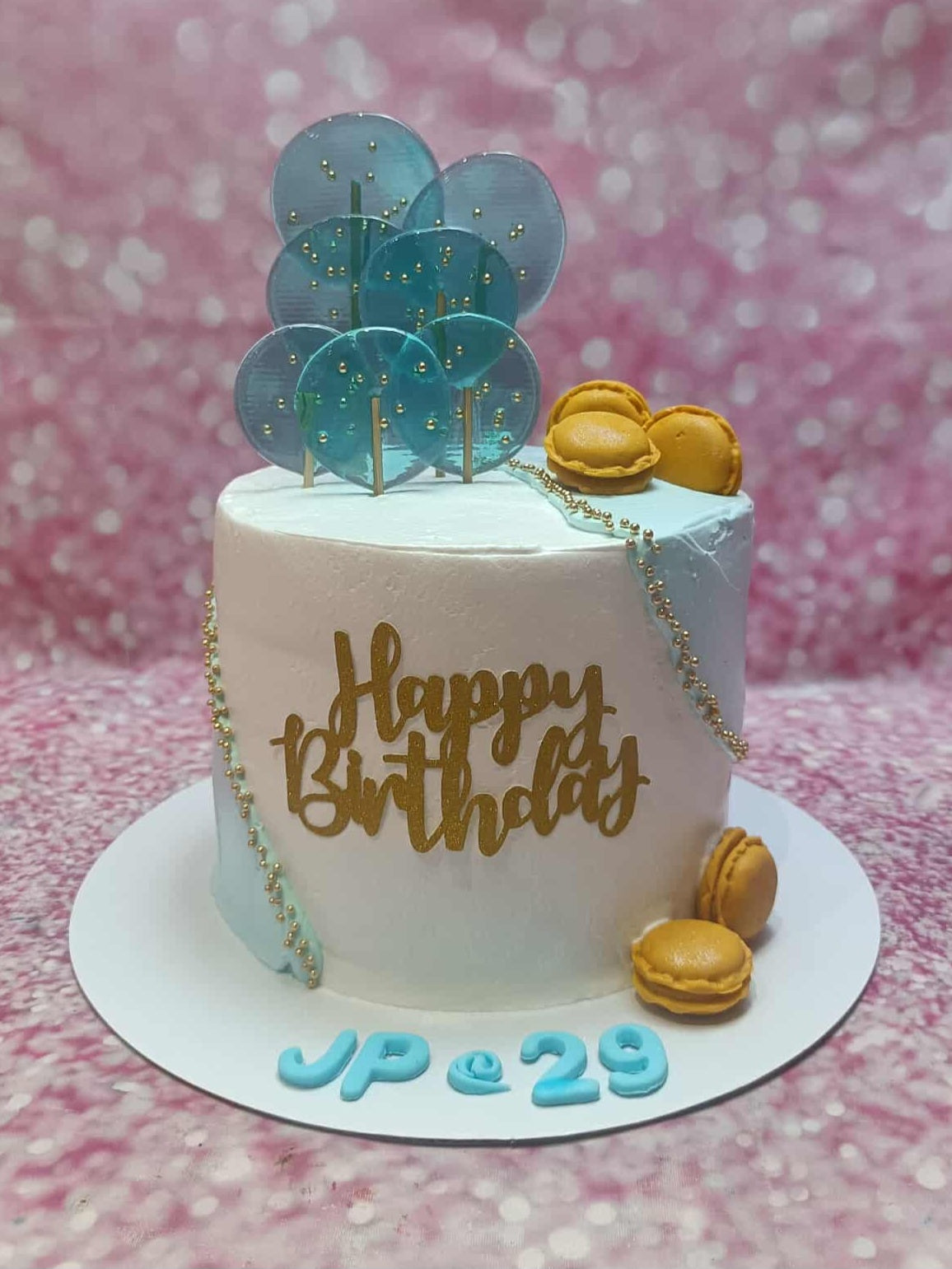 One-tier birthday cake with white & blue buttercream, gold "Happy Birthday" topper, teal lollipops, macarons, "JP @ 29".