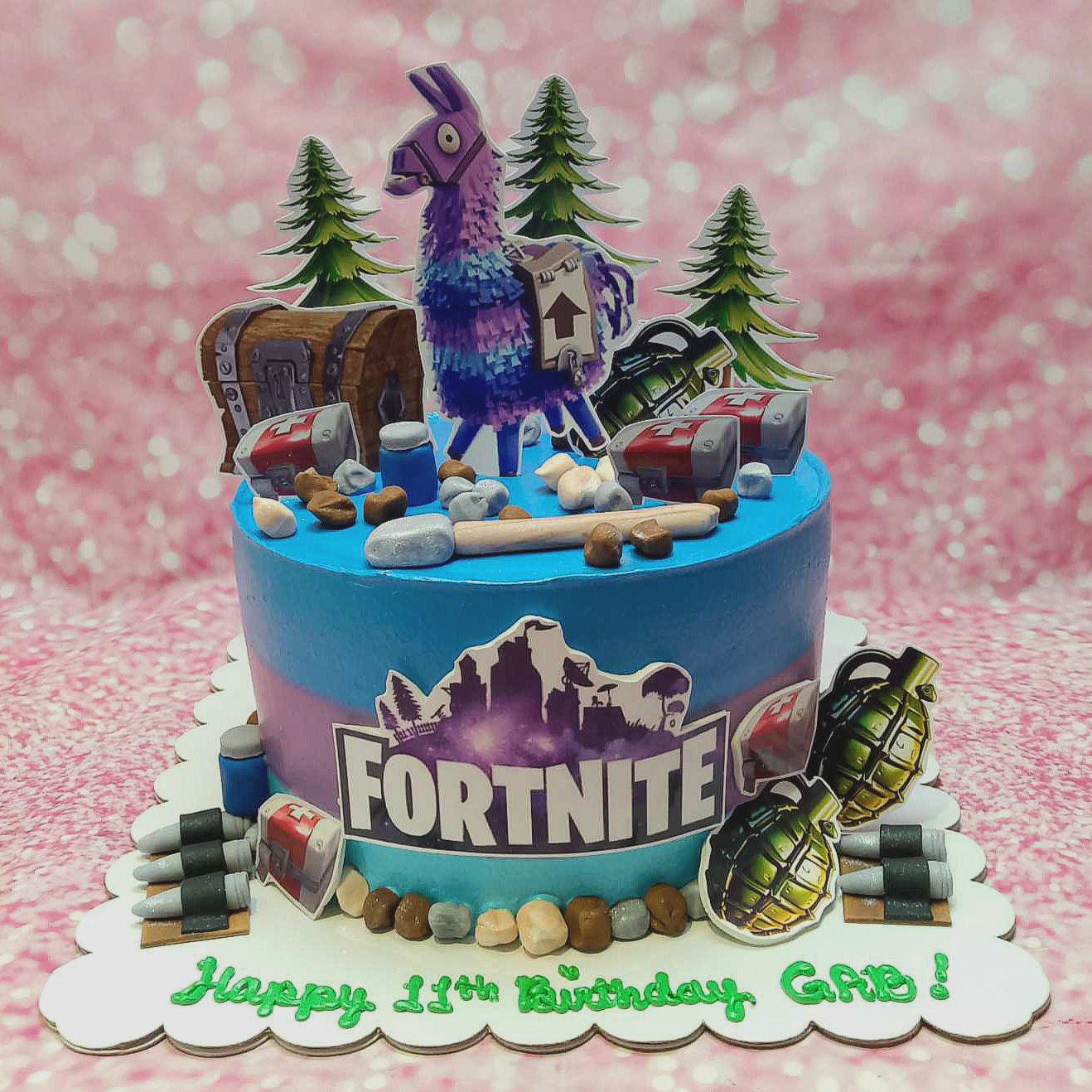 This Fortnite Cake features a stunning blue and purple ombre design, adorned with a playful Llama, trees, chests, grenades, bullets, and rocks, perfect for celebrating Gab's 11th birthday. The cake is topped with vibrant printout toppers inspired by video games on PS5 and Xbox, showcasing a single tier for a unique party centerpiece.