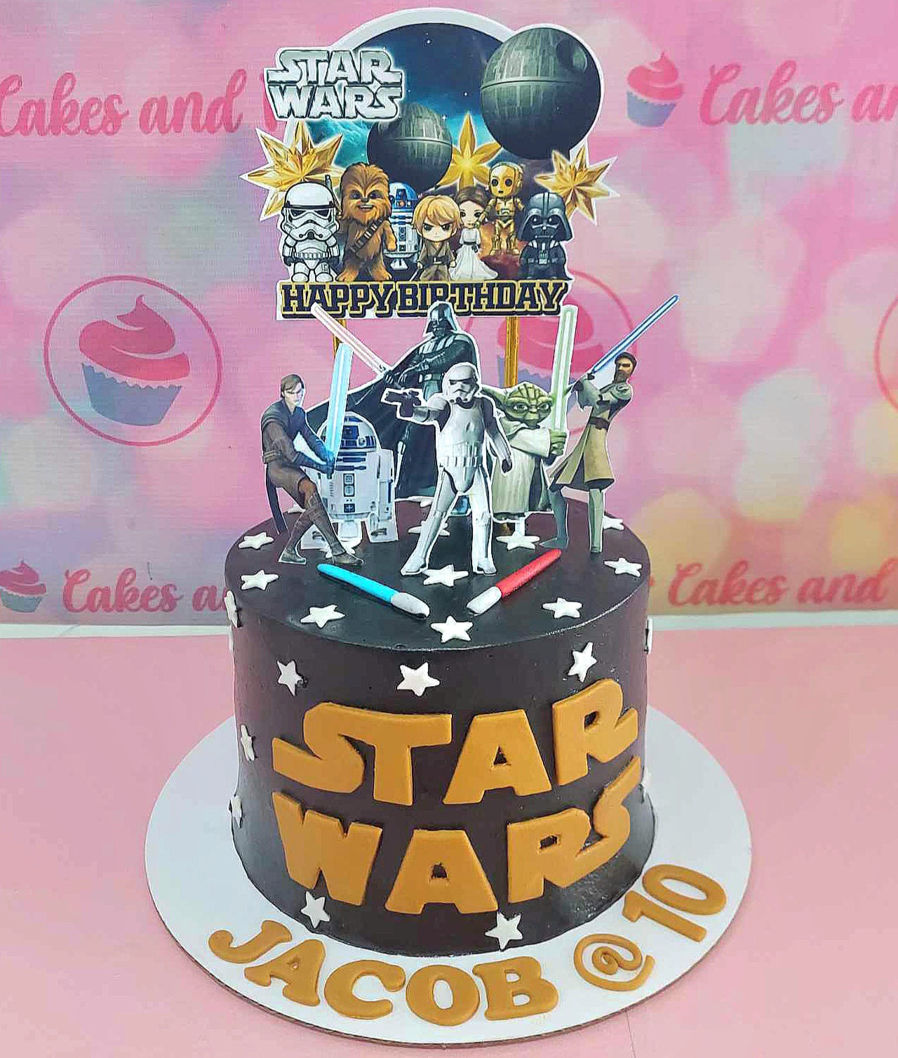 This custom Star Wars cake features a striking black and gold design, adorned with themed printout toppers of characters like Darth Vader, Yoda, and Chewbacca, perfect for a 10th birthday celebration. The cake is topped with lightsaber accents and a decorative Death Star, capturing the essence of the galaxy far, far away for any young Jedi fan.