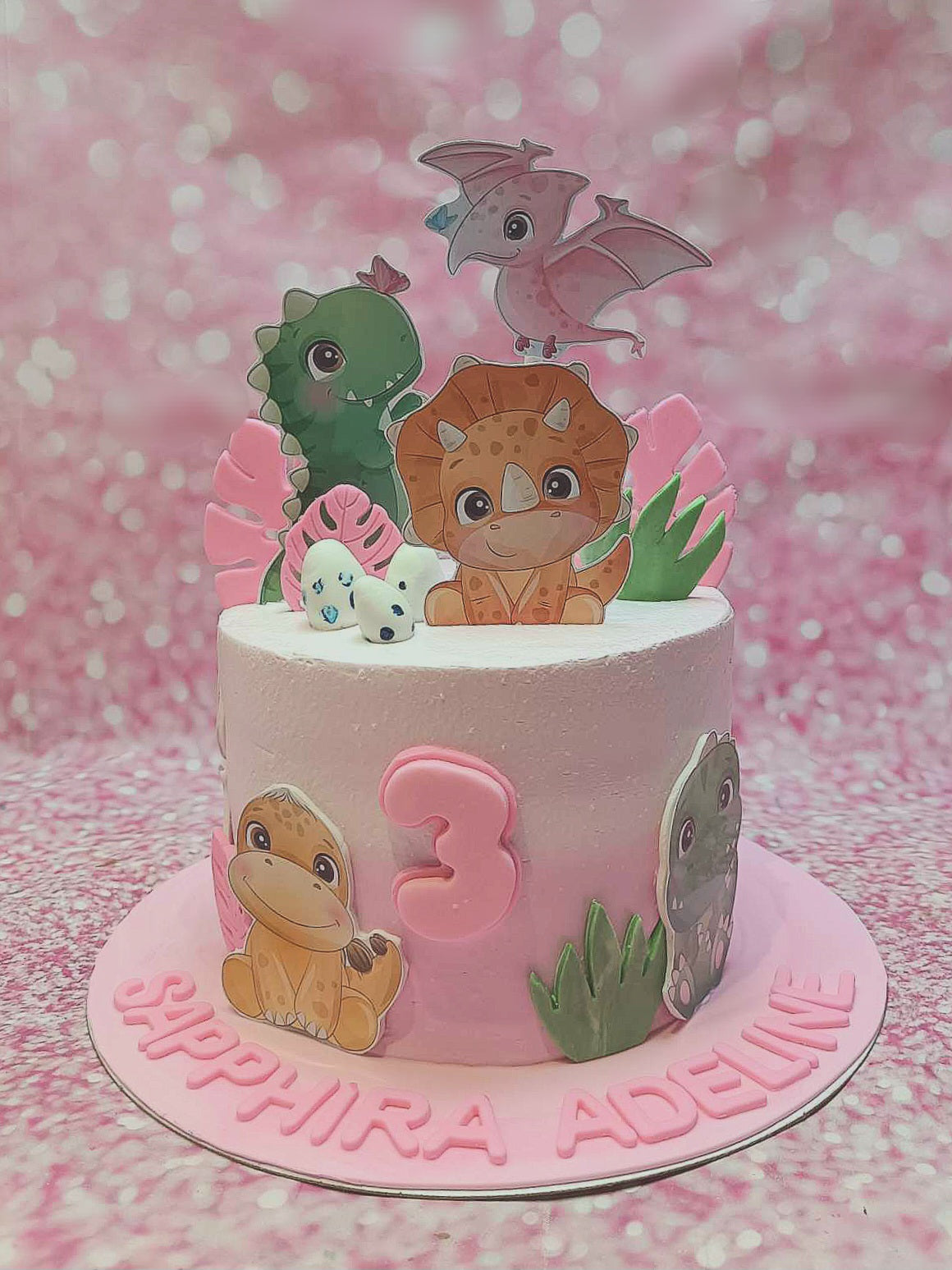 This charming dinosaur cake is adorned with pink icing and features adorable dinosaur printout toppers, perfect for celebrating Sapphira Adeline's 3rd birthday. Nestled among colorful eggs and leaves, this one-tier cake captures a delightful dinosaur theme.