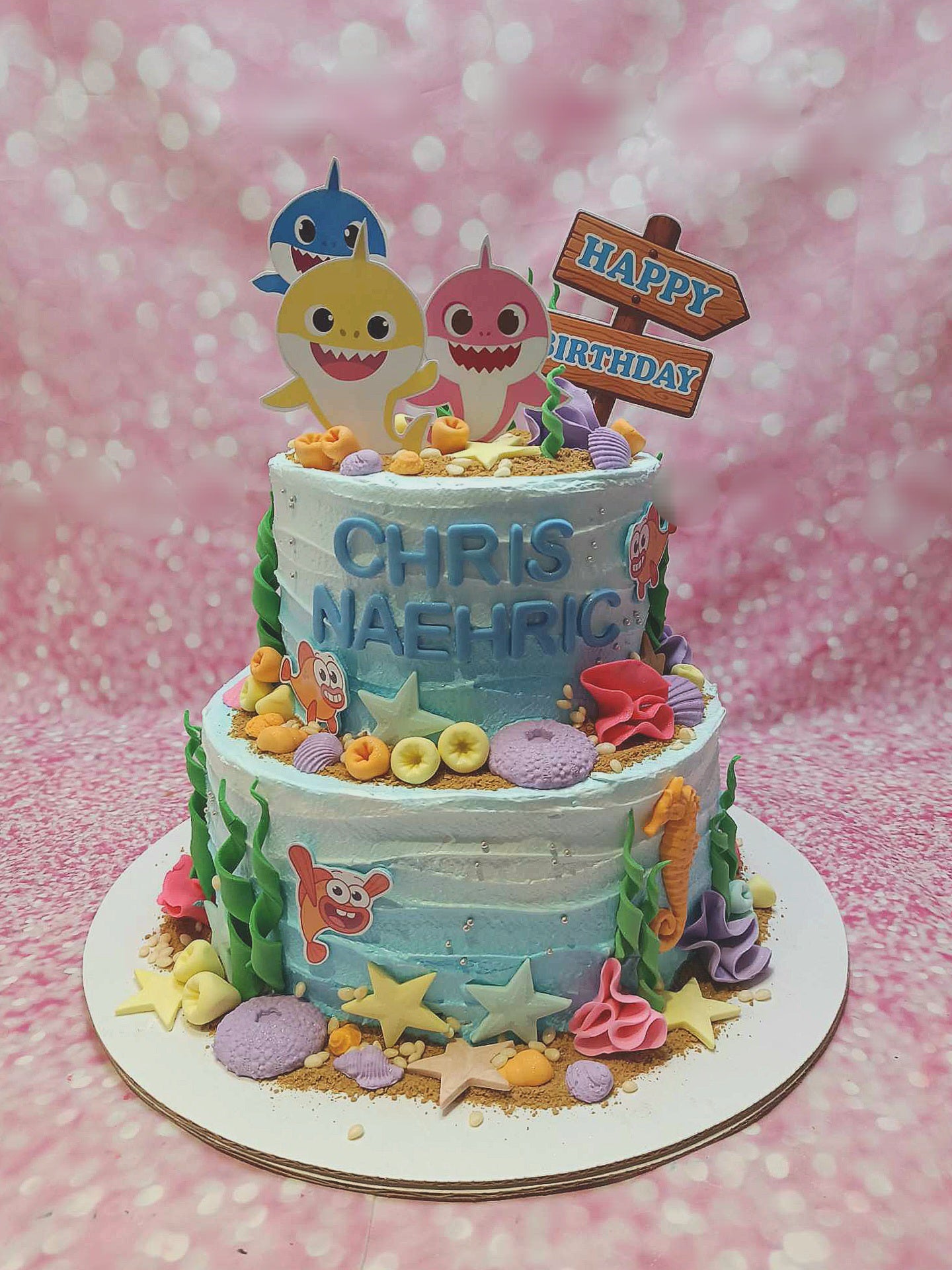This stunning two-tiered Baby Shark cake features light blue icing adorned with playful printout toppers of Baby Shark, starfish, seashells, seahorse, and coral, making it a perfect centerpiece for a birthday celebration. A cheerful "Happy Birthday" sign completes the design, showcasing the creativity of Chris Naehring.