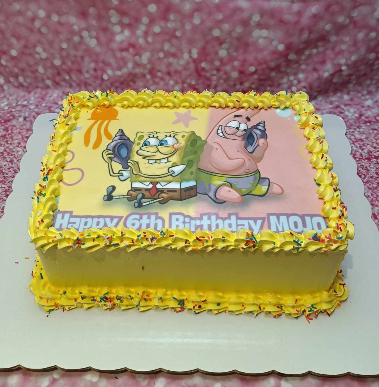 This custom Spongebob Cake celebrates Mojo's 6th birthday with a colorful design featuring an edible photographic print of SpongeBob Squarepants and Patrick Star, adorned with shells, starfish, and sprinkles in shades of yellow and pink. The rectangular cake is a perfect centerpiece for any themed celebration.