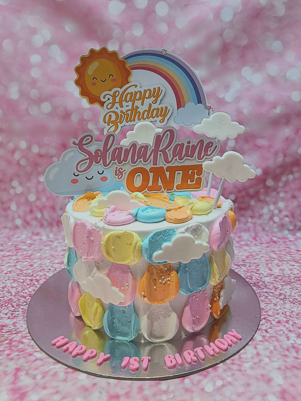 The Sunshine Cake features pastel colors and delightful decorations, including sun and rainbow motifs surrounded by fluffy cloud toppers. Perfect for a 1st birthday celebration, this charming 1-layer cake radiates happiness and captures the essence of a sunny summer morning.
