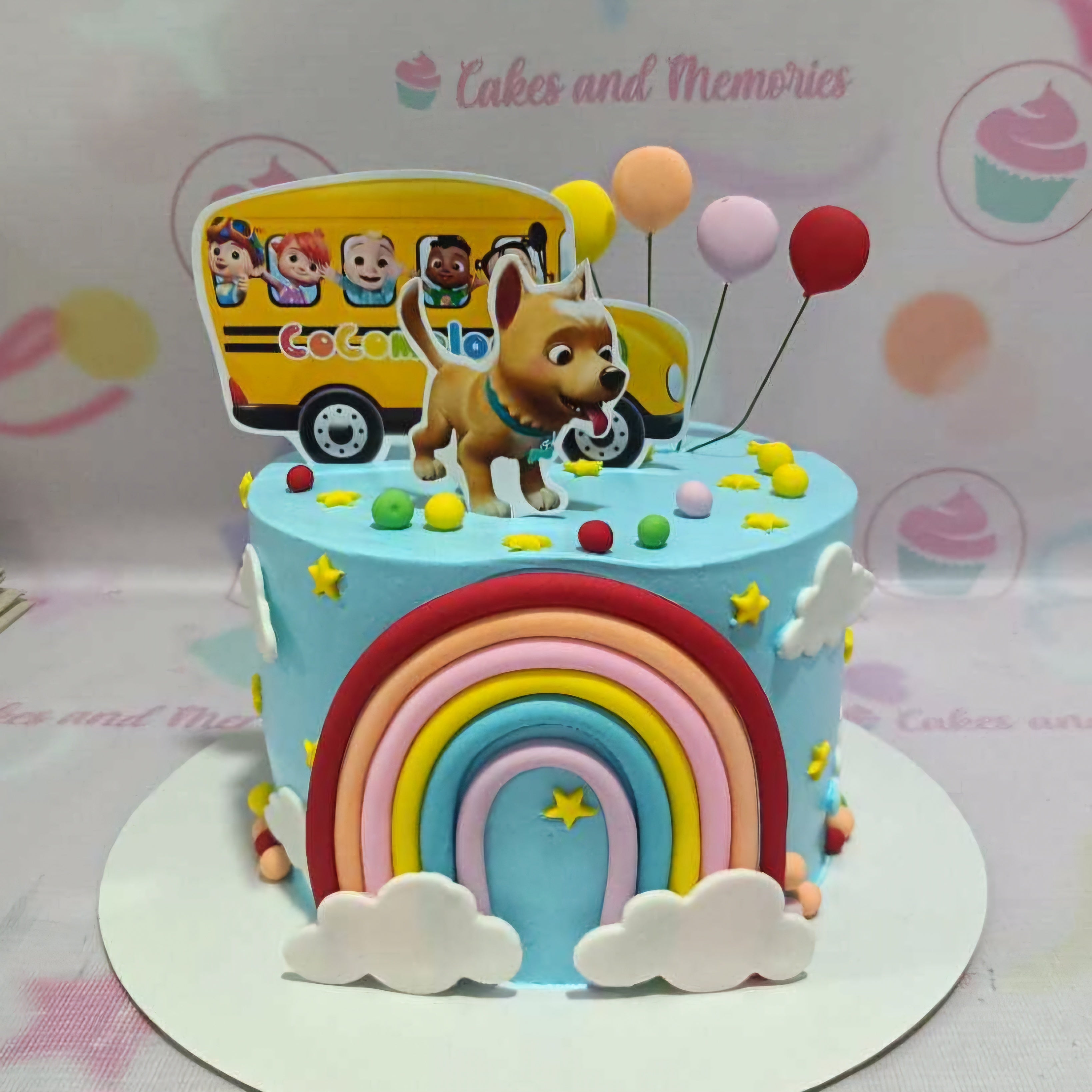 This vibrant Cocomelon cake features a playful blue design with a cheerful yellow bus, colorful balloons, and an adorable dog, perfect for a baby's first birthday celebration. The customized printout toppers highlight the "Wheels on the Bus" theme, creating a fun and festive centerpiece.