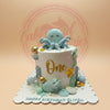 Under the sea first birthday cake with a light blue octopus, turtle, seahorse, gold 'One', and 'Eliyah' on a white board.