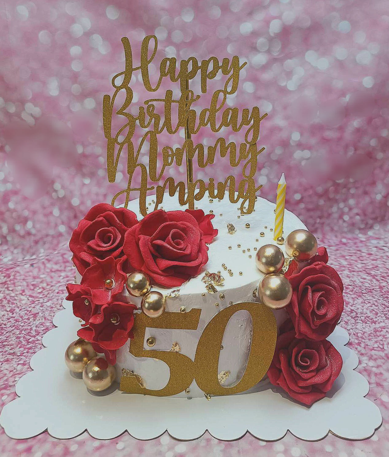 This elegant cake features smooth white icing adorned with red roses and edible pearls, perfect for celebrating milestones like a 50th birthday. Topped with a stunning gold glitter cardstock "50" and personalized for Mama, it's a beautiful centerpiece for any family gathering.