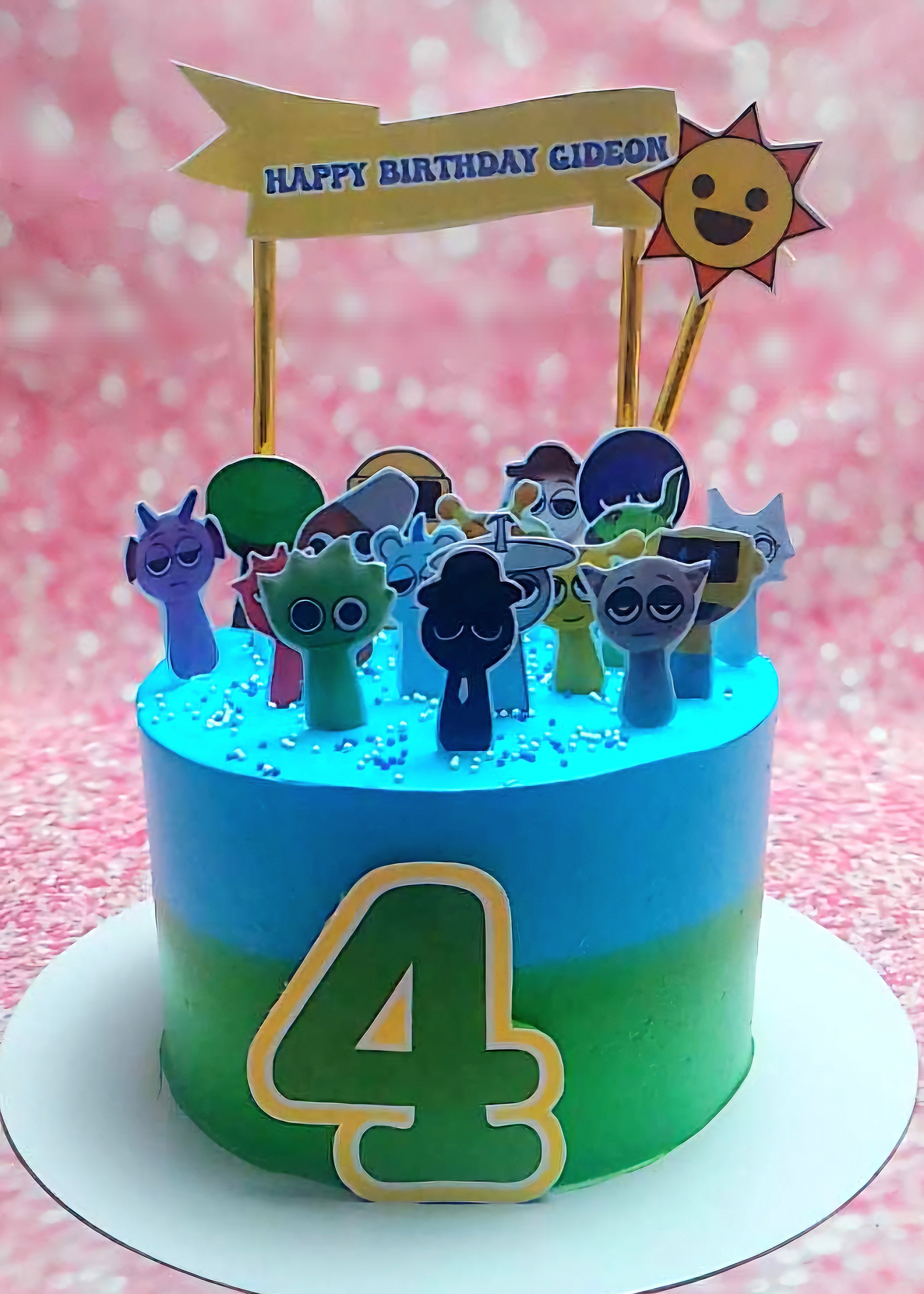 This eye-catching birthday cake features a blue and green ombre design adorned with sprunky cartoon characters and a sun, perfect for a 4th birthday celebration. Topped with a yellow banner and printout toppers from popular horror video games, it's a fun centerpiece for the birthday boy's special day.