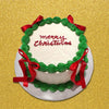 This elegant Christmas cake features a smooth white surface adorned with a festive wreath design, red ribbon bows, and green swirls, perfect for any Christmas party or celebration. The single tier showcases a cheerful 