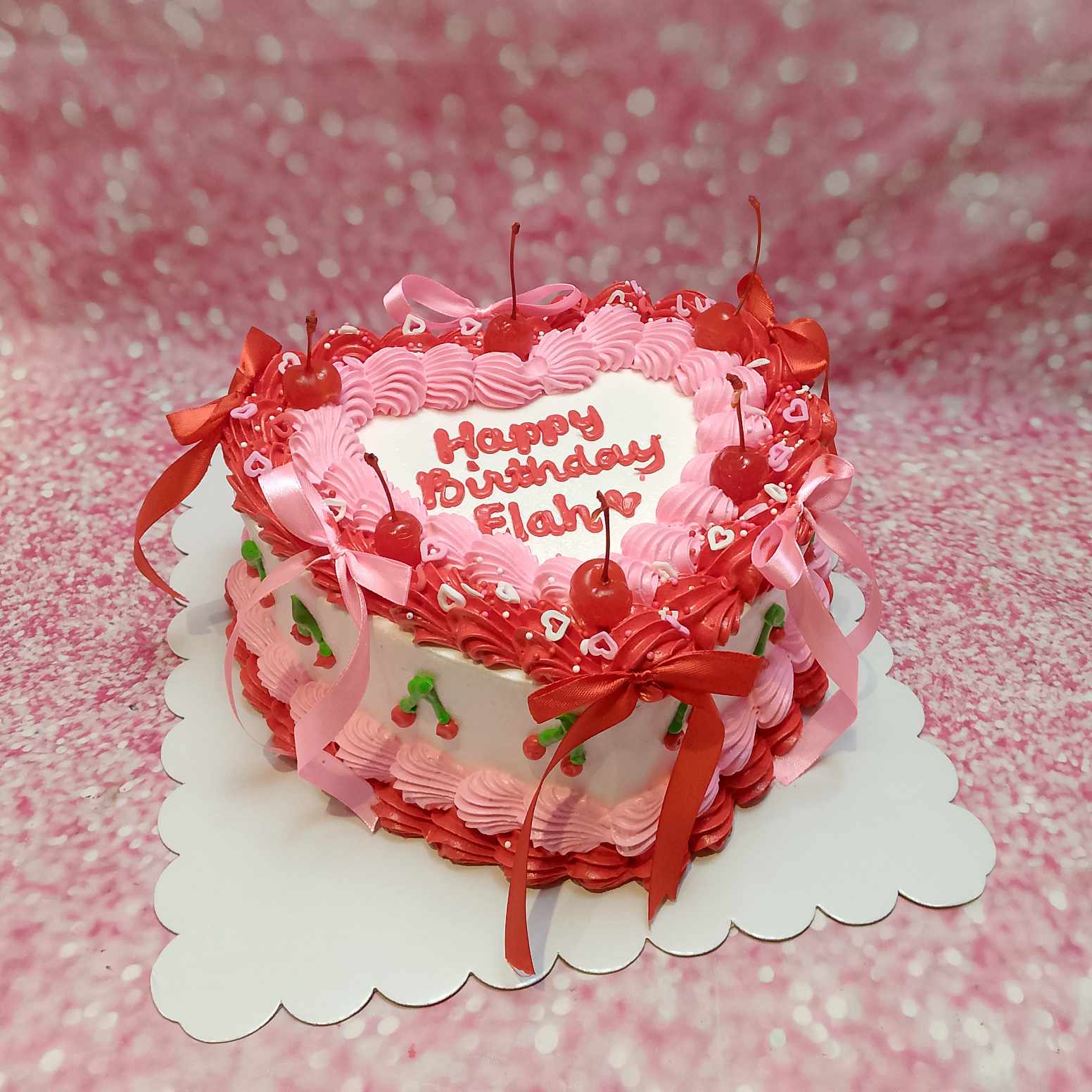 This vintage pink and red heart-shaped cake features a charming ribbon design adorned with cherries, bows, and heart sprinkles. Perfect for any celebration, it showcases a delightful one-tier style with a nostalgic touch.