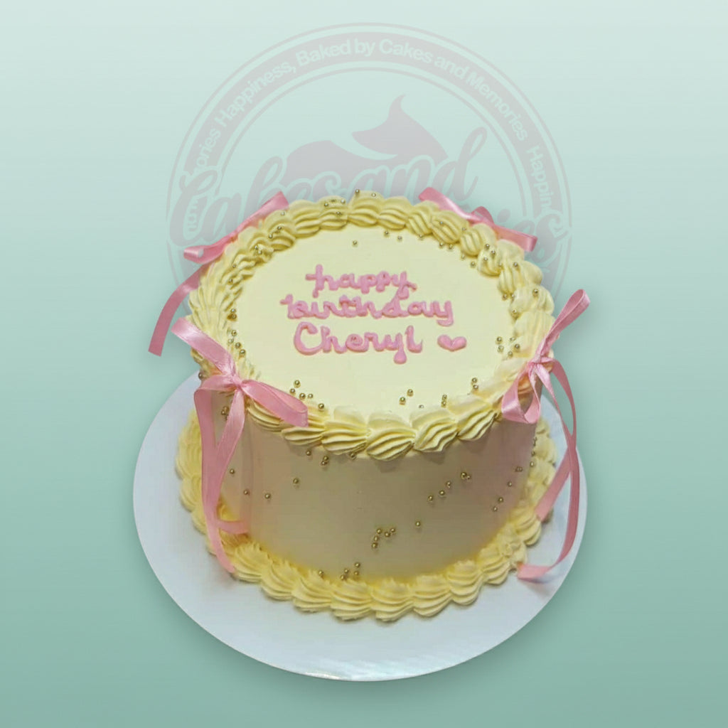 Pale yellow buttercream birthday cake with pink hand-piped 'happy birthday Cheryl!', gold pearls, and pink satin ribbons.