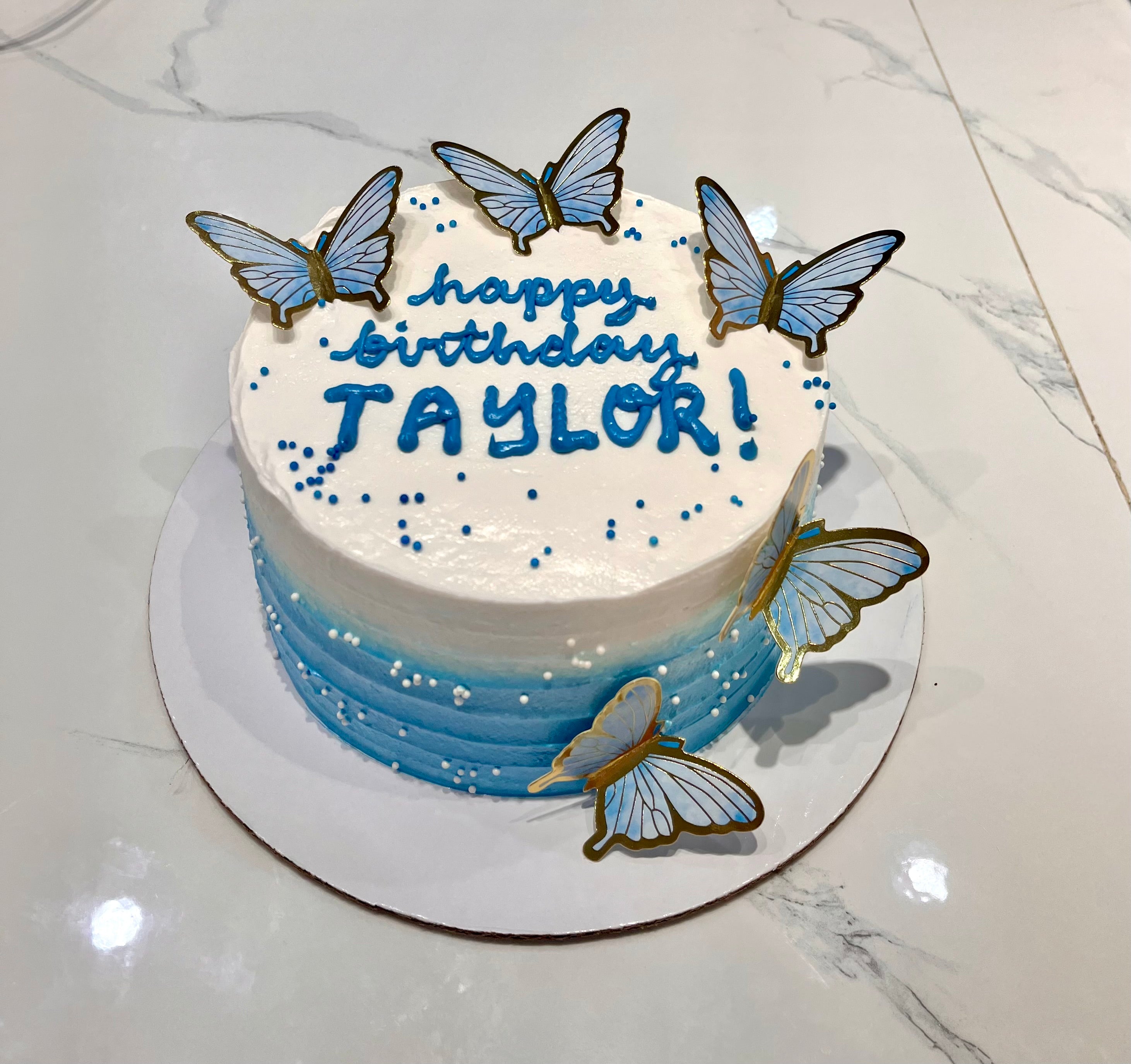 This Butterflies Cake features a stunning blue ombre design with delicate white butterflies and colorful sprinkles. The minimalist design is complemented by unique printout toppers, showcasing an elegant single layer and tier.