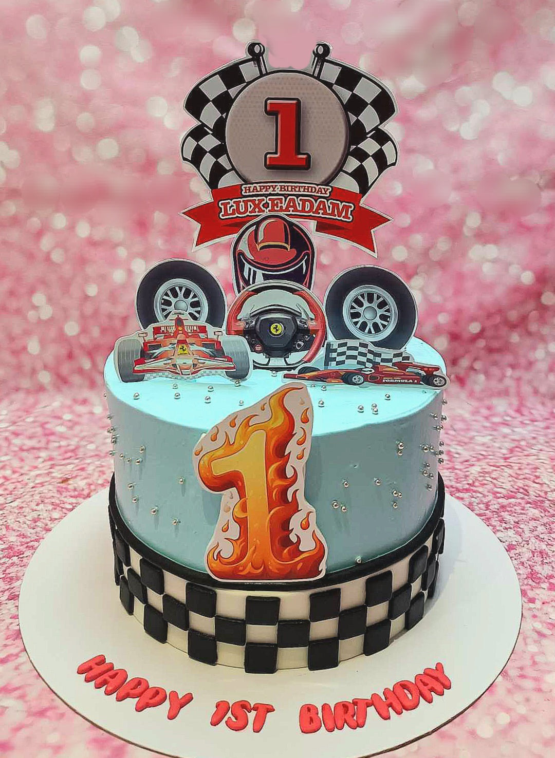 This light blue racing cake features a sleek design with a black and white checkered pattern, perfect for a 1st birthday boy. Adorned with race car and Ferrari printout toppers, it showcases a fun racing theme with a steering wheel accent.