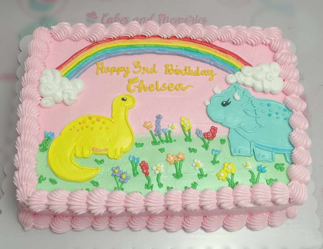 This custom Dinosaurs Cake features a playful pink color scheme with fun doodle designs, including clouds and flowers, perfect for a girl's 3rd birthday. The rectangular cake showcases adorable triceratops among a colorful rainbow backdrop.