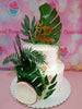 This elegant two-tier Garden Cake features lush green leaves and delicate flowers, adorned with gold leaf accents for a modern touch. Crafted in a coconut shell design, it beautifully complements the tropical theme with its simple yet striking layers.