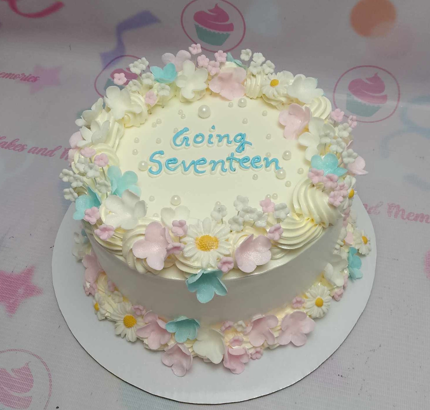 This one-tier Going Seventeen Cake features a white base adorned with pastel blue and pink daisies, perfect for a 17th birthday celebration. Completed with charming printout toppers, it captures the essence of a garden party with a cute K-pop flair.